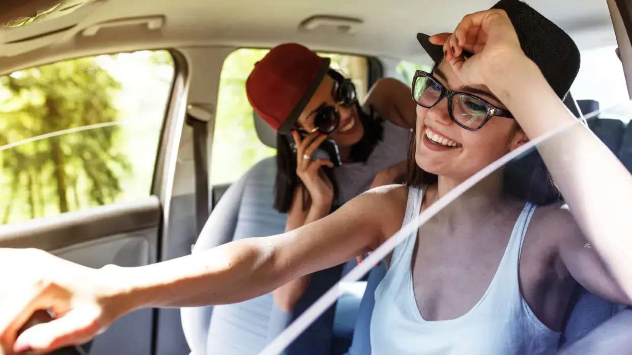 Duas mulheres sorridentes dentro de um carro, uma ao volante e outra no banco de trás a falar ao telemóvel.