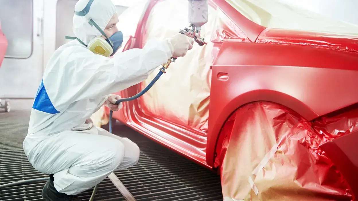 Trabalhador com equipamento de proteção a pintar carro vermelho numa cabine de pintura