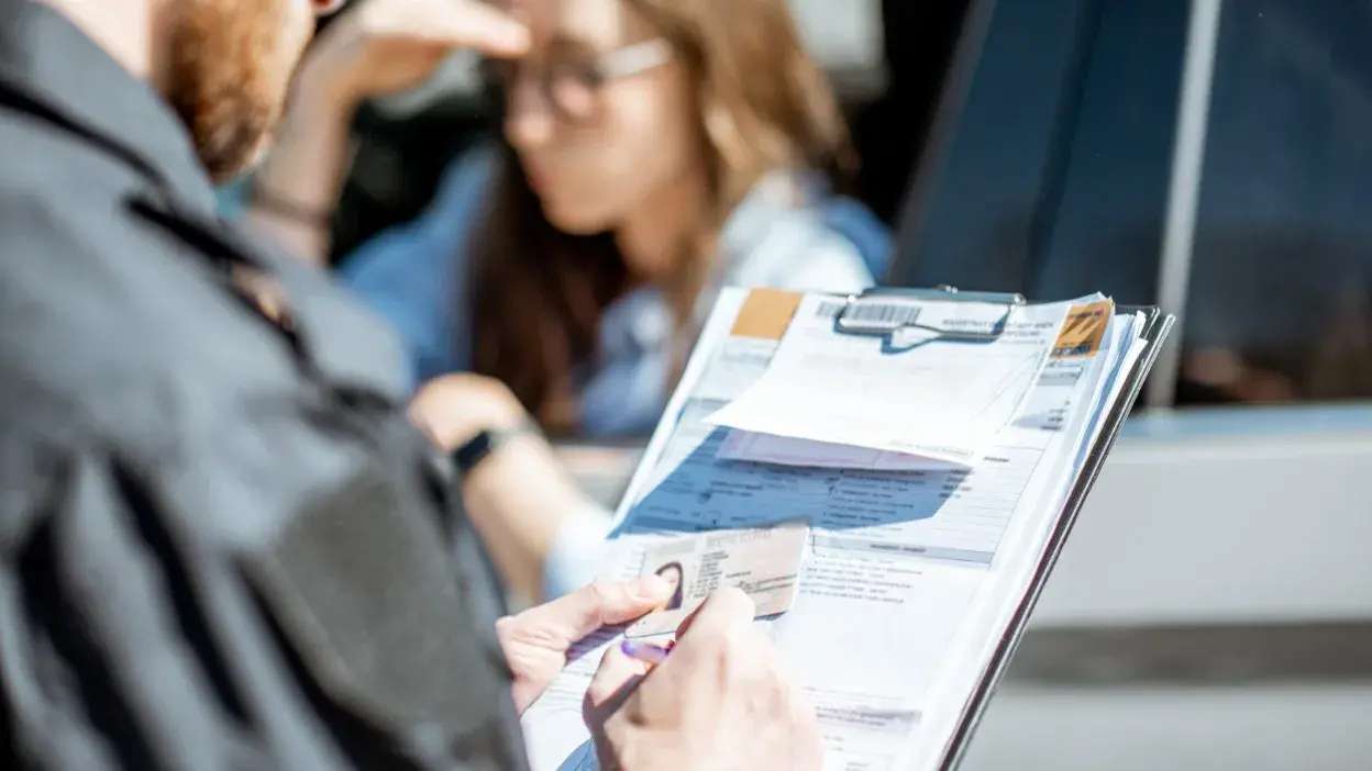 Pessoa a mostrar documentos e multa de trânsito junto a um carro com outra pessoa ao fundo