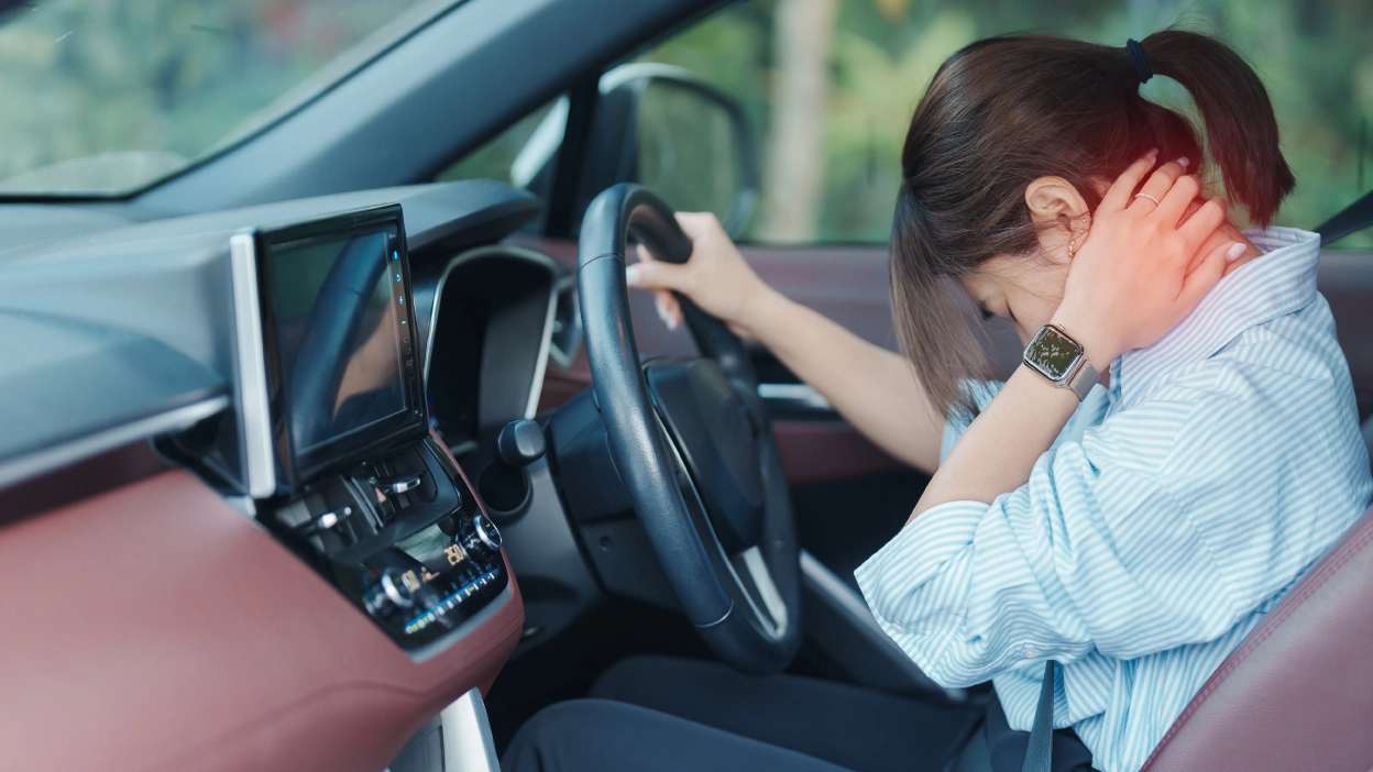 mulher sentada dentro do carro, visivelmente cansada, com a cabeça para baixo e mão no pescoço