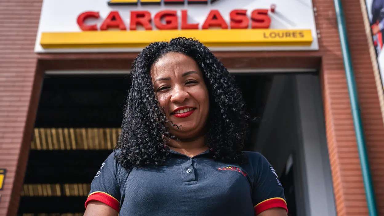 Colaboradora da Carglass em frente à oficina da marca, com uniforme profissional e ambiente exterior da oficina automóvel. 